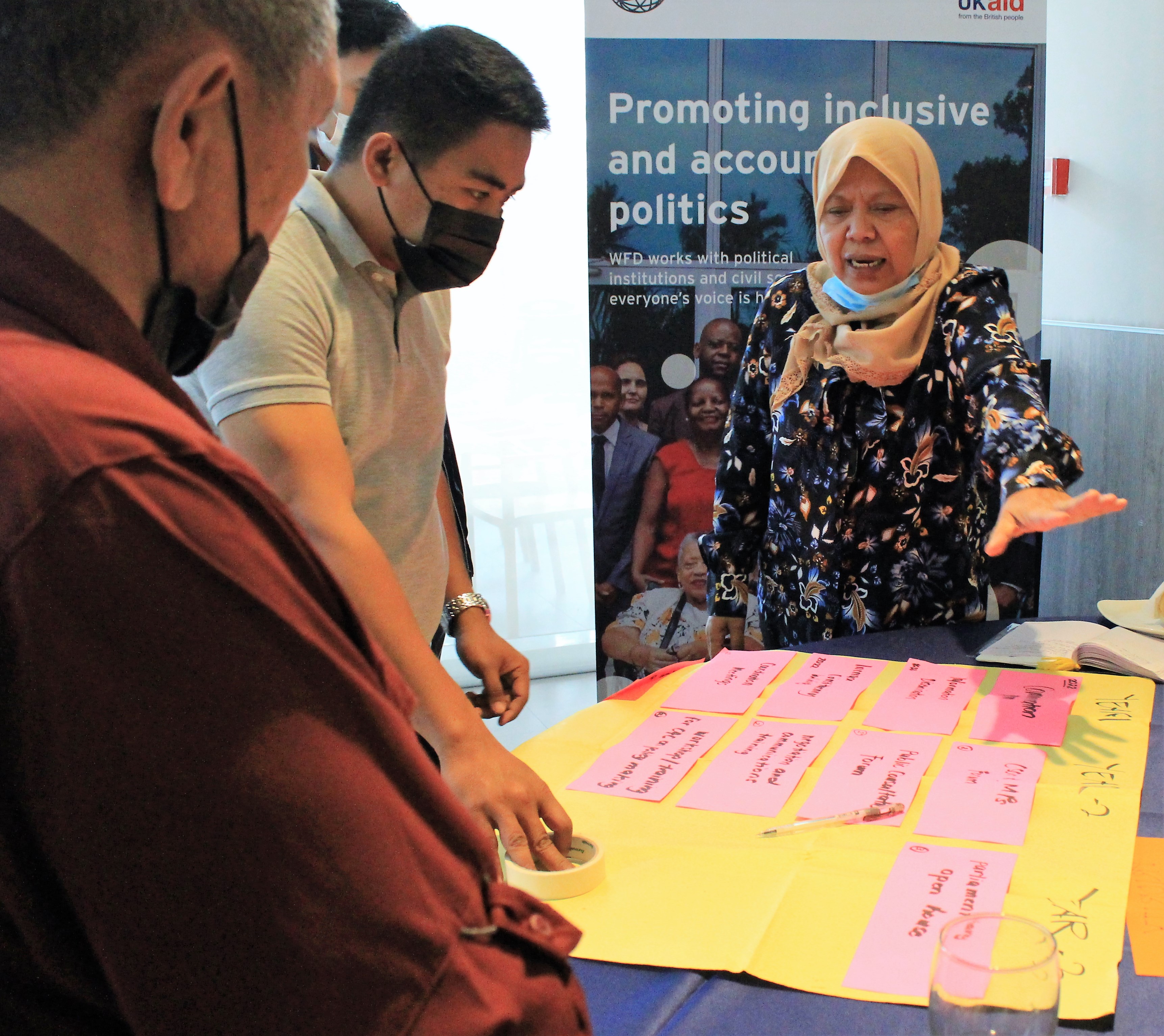 A woman wearing a headscarf stands speaking in front of a table of post-it-notes. Two men in face masks listen with concentrated expressions