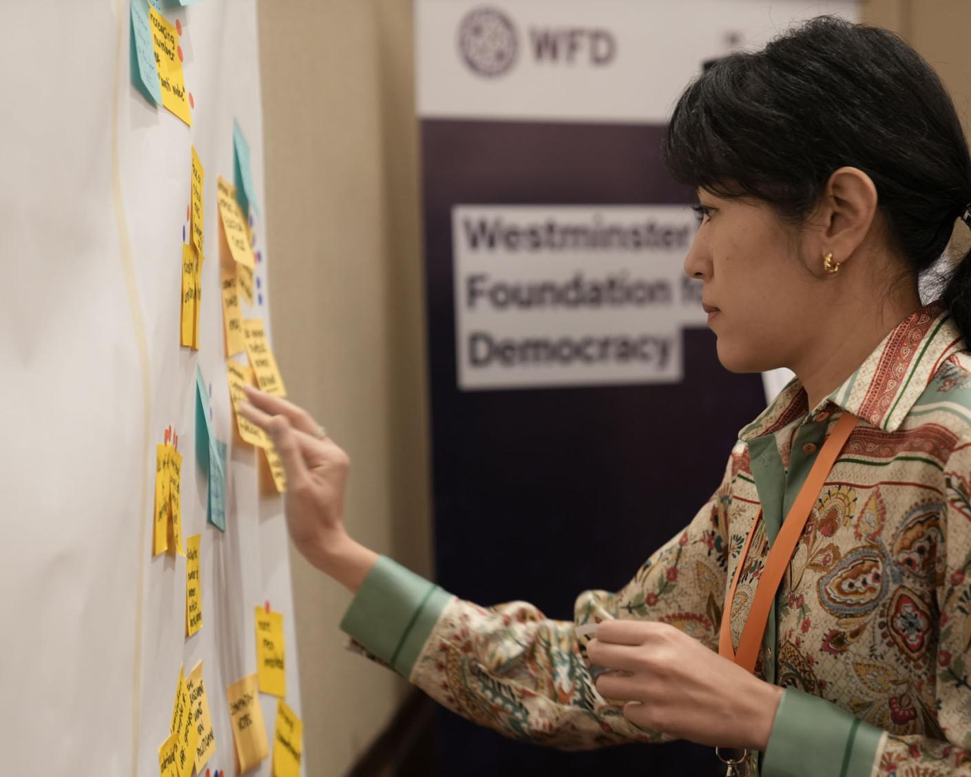 A woman adds her contribution on a sticky note to a wall of other notes. She stands in front of a sign on which the words Westminster Foundation for Democracy can be read