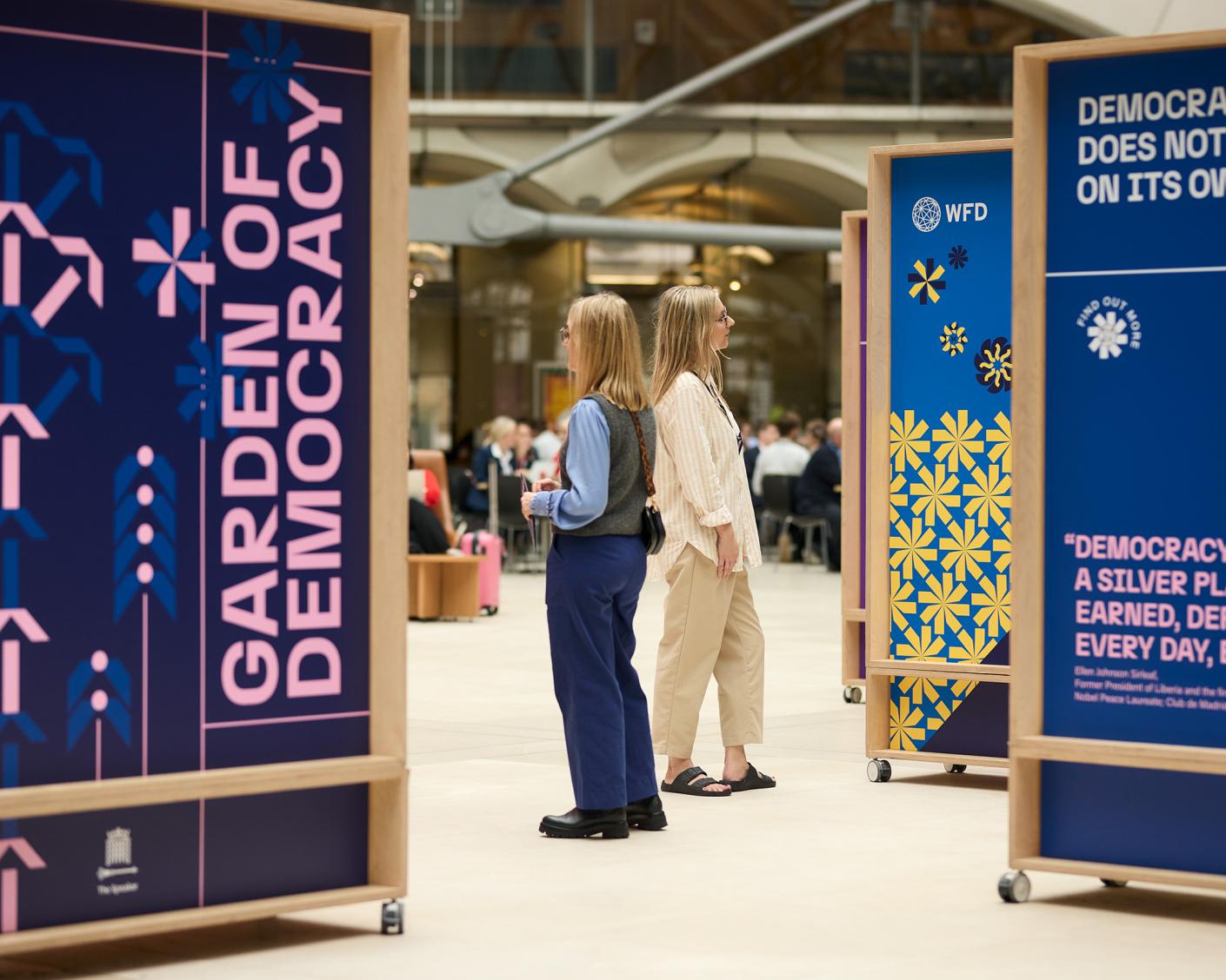 Two women stand between exhibition panels, one of which reads "garden of democracy"
