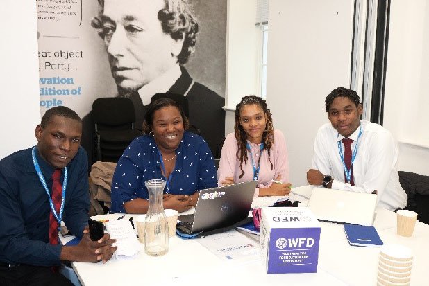 Image of four people sitting at a table gathered around a laptop, and smiling at the camera