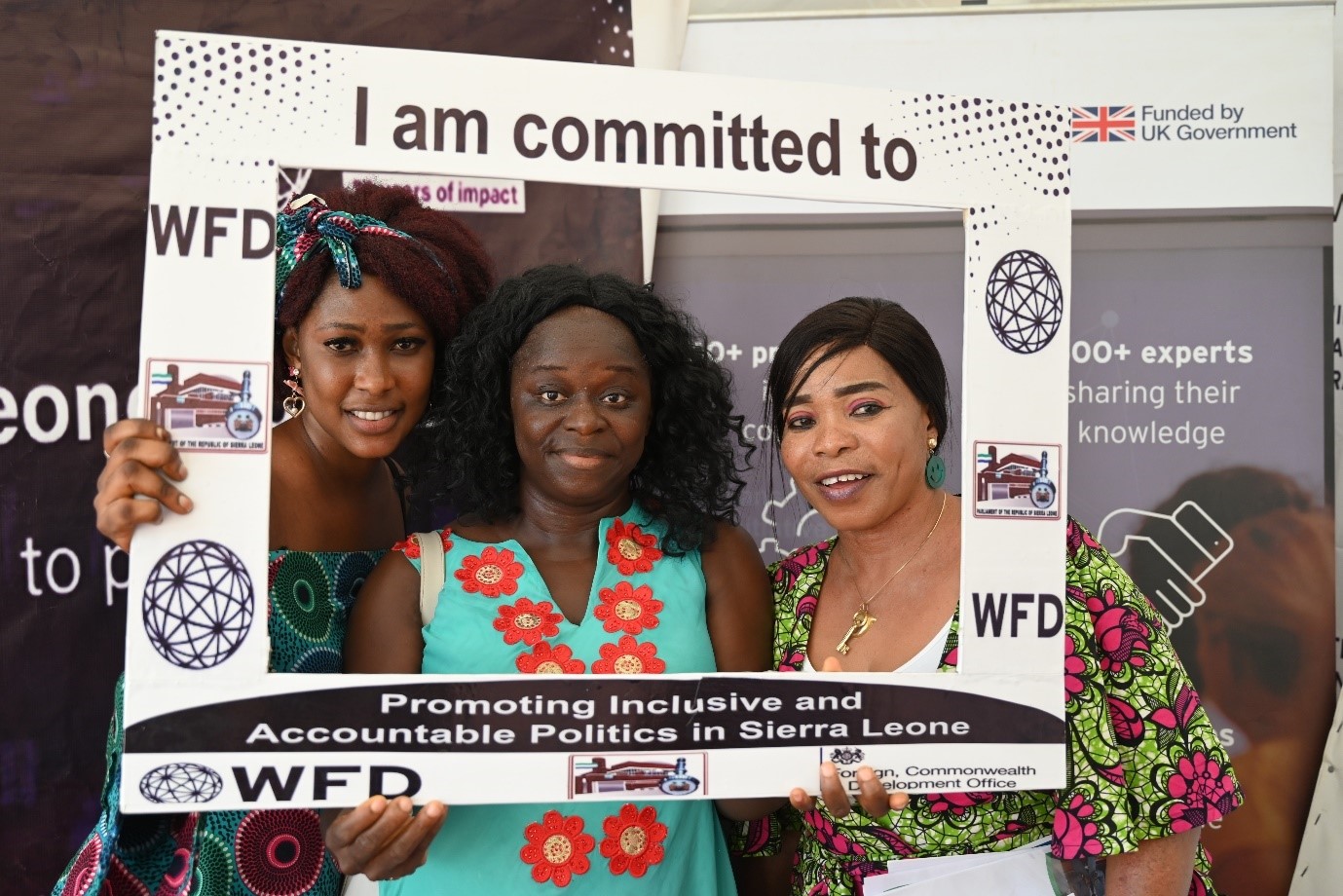 Three women holding up a sign that says I am committed to promoting inclusive and accountable politics in Sierra Leone