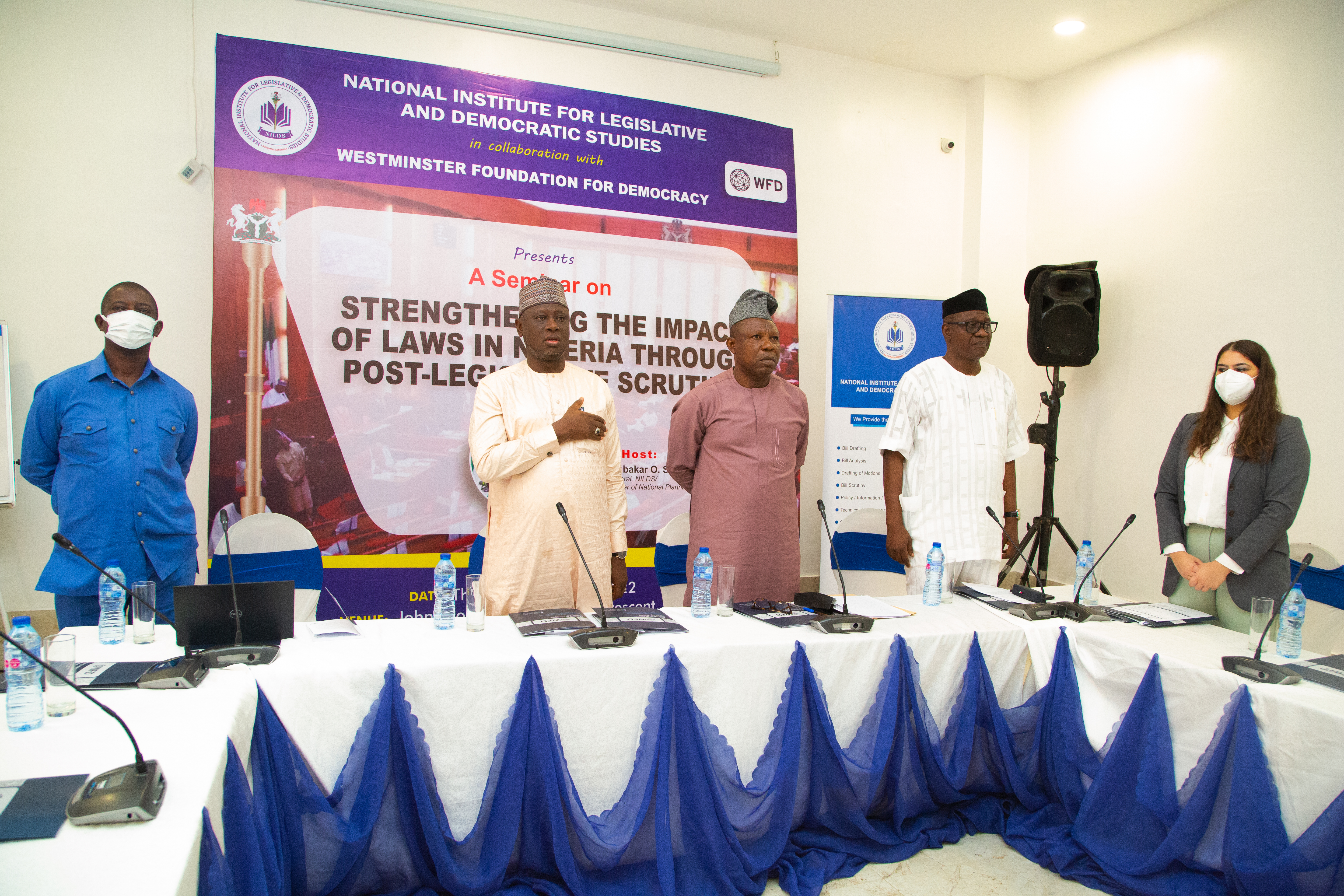 Several men standing in front of a poster advertising a PLS conference in Nigeria. One has his hand on his heart.