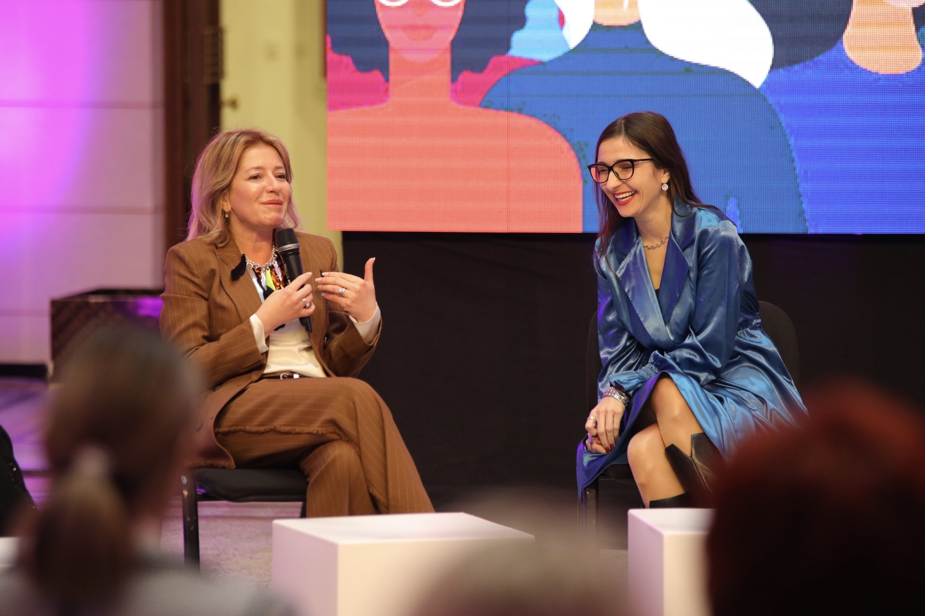 Two women speaking on a stage. One is using a microphone and the other is listening. Both are smiling.