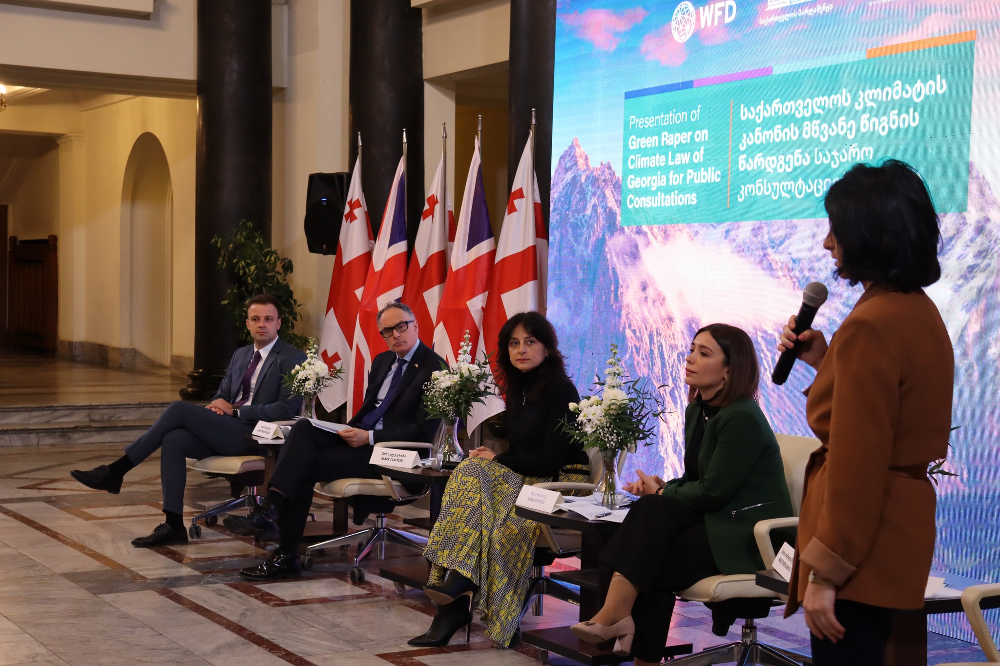 A woman speaks into a microphone at a panel event. In the background are an image of mountains and Georgian flags
