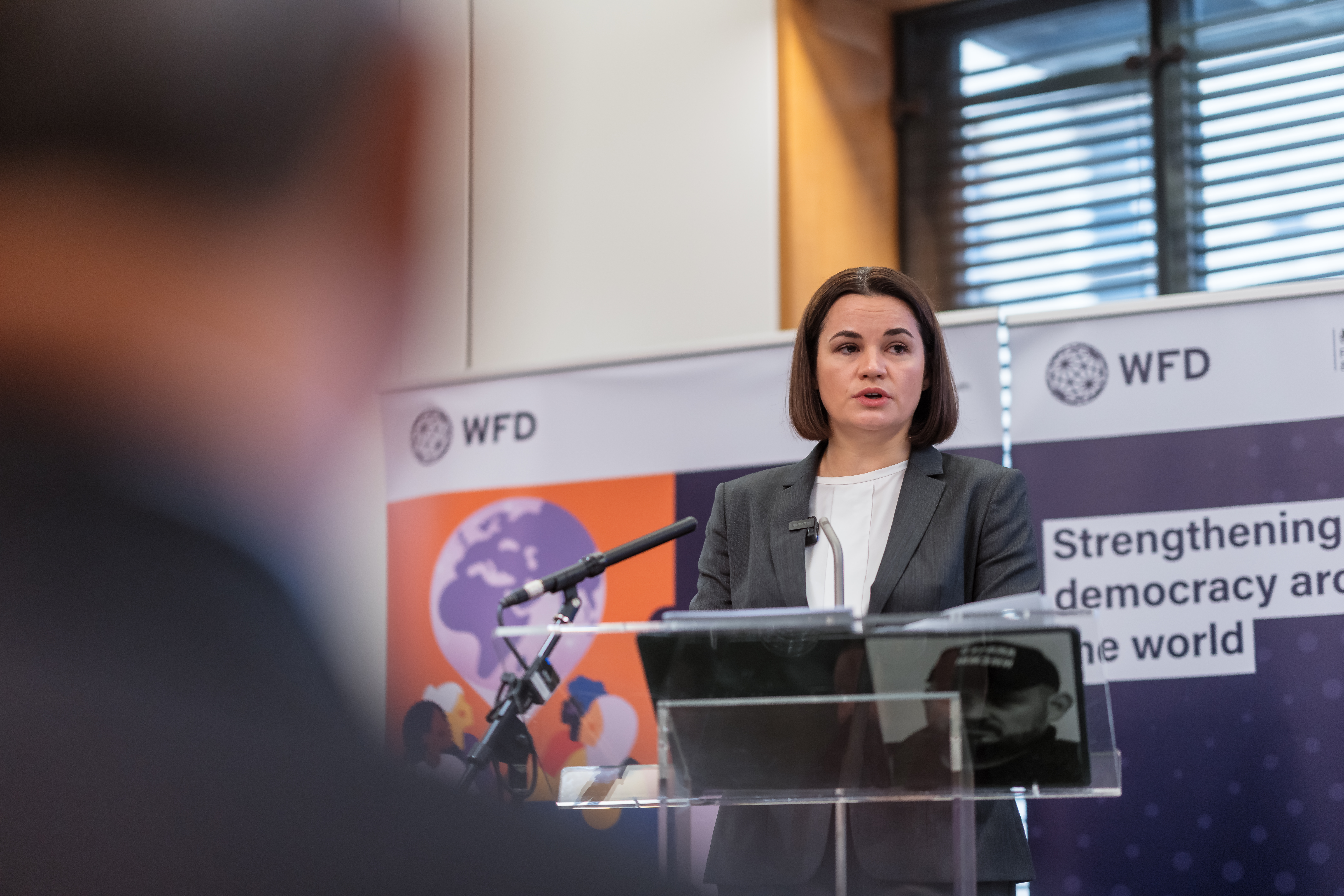 A woman stands at a lectern in front of a microphone, the backs of the audience's heads are in the foreground. She stands next to a sign that says strengthening democracy around the world. The words Inaugural Lecture on the State of Democracy are on a screen next to her.