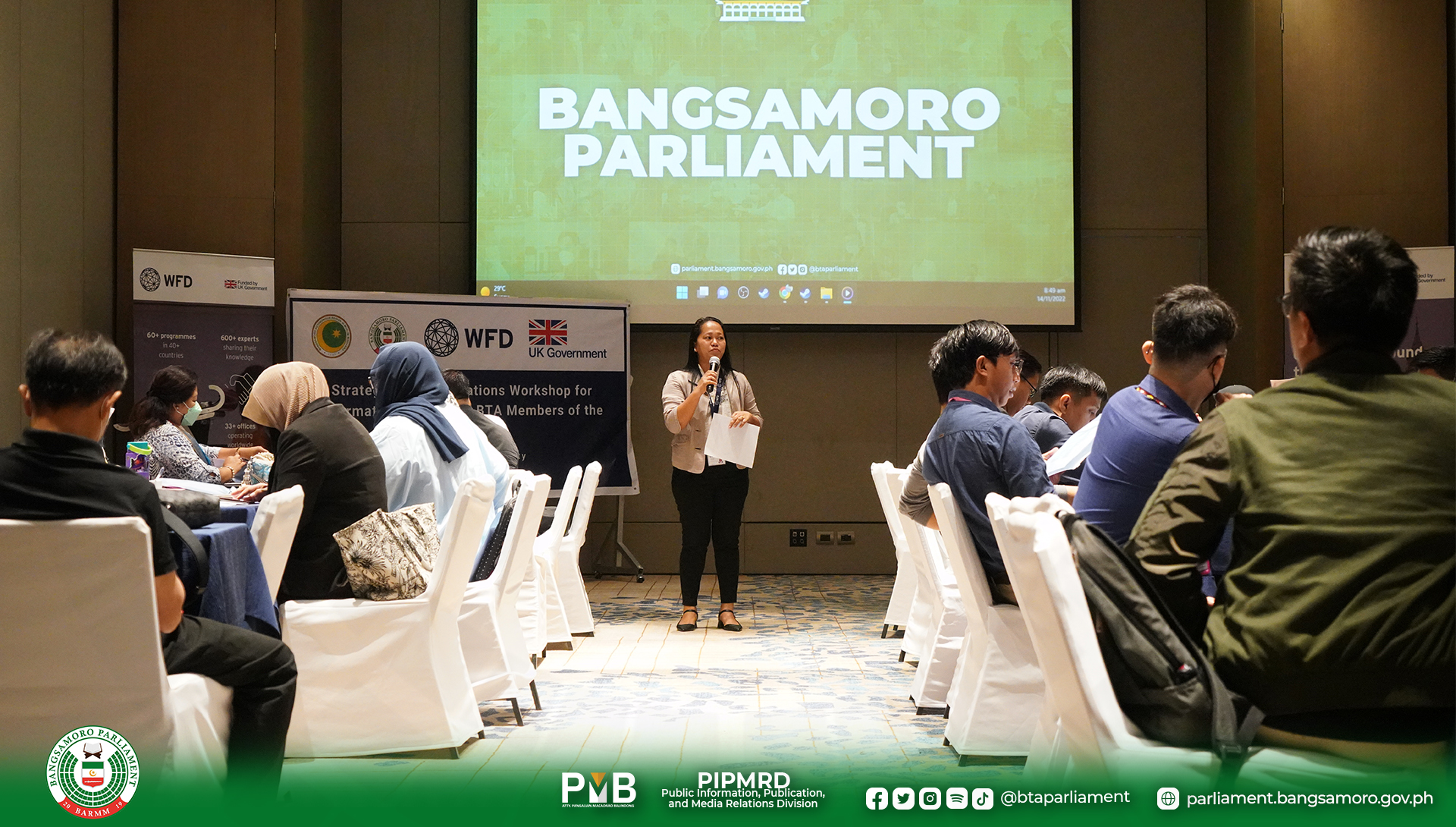 A woman with a microphone stands in front of an audience. Behind her a screen says Bangsamoro Parliament