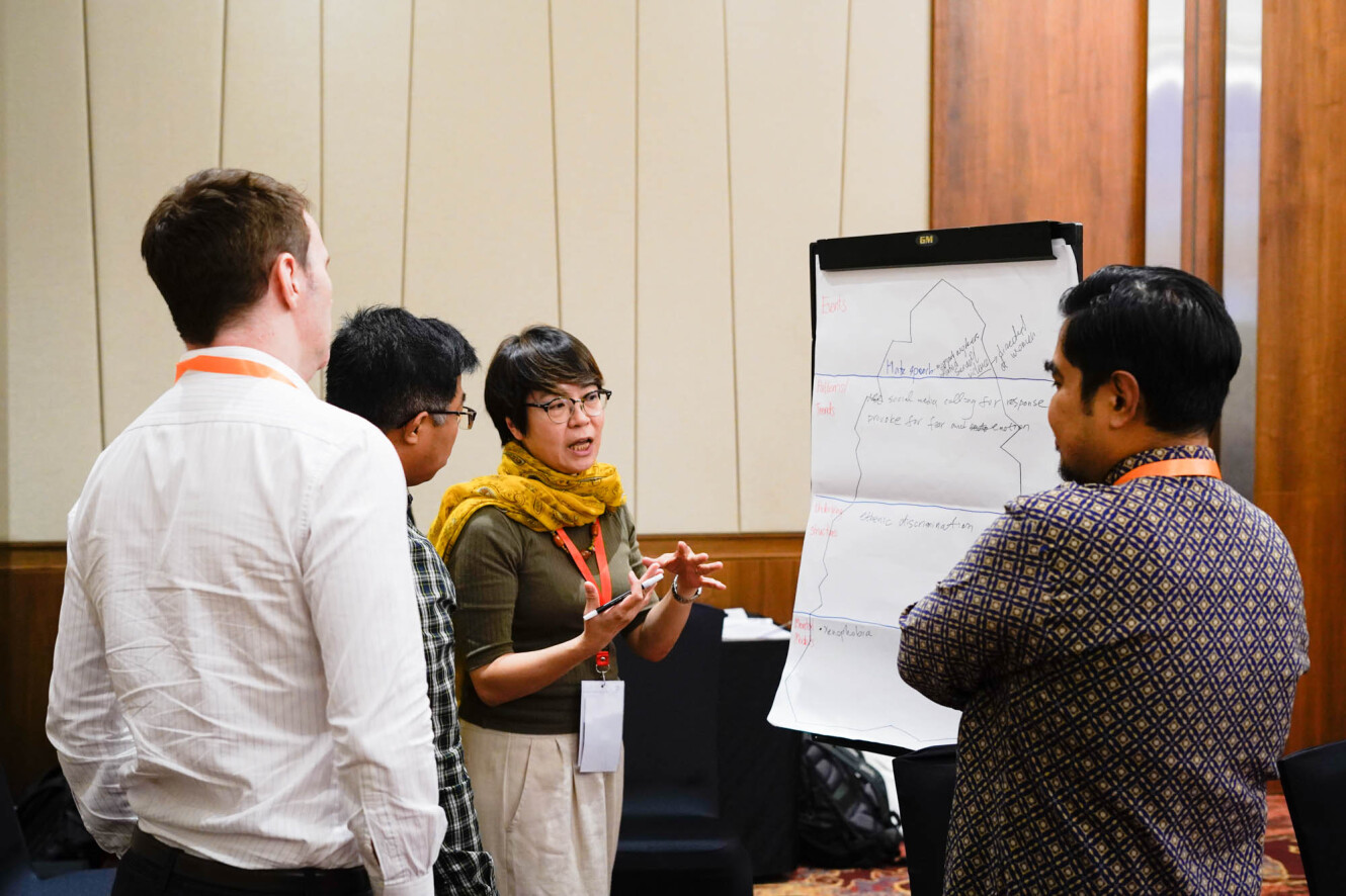 A group of people stand around a flipchat, they are listening to a woman who gesticulates