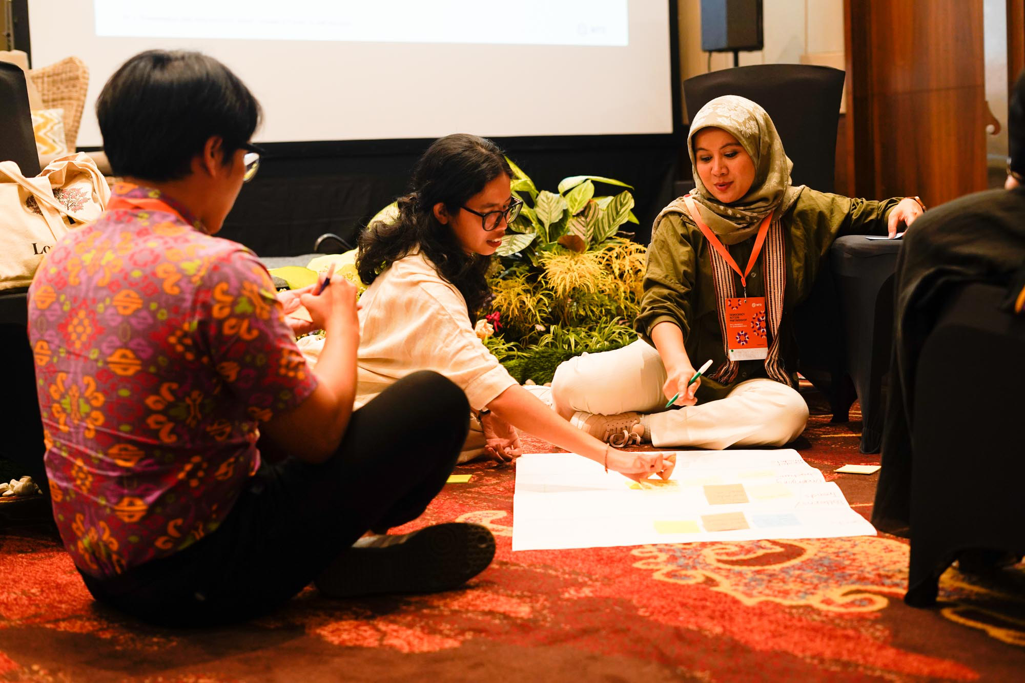 Three people sit round a large piece of paper adding post-it notes and writing notes