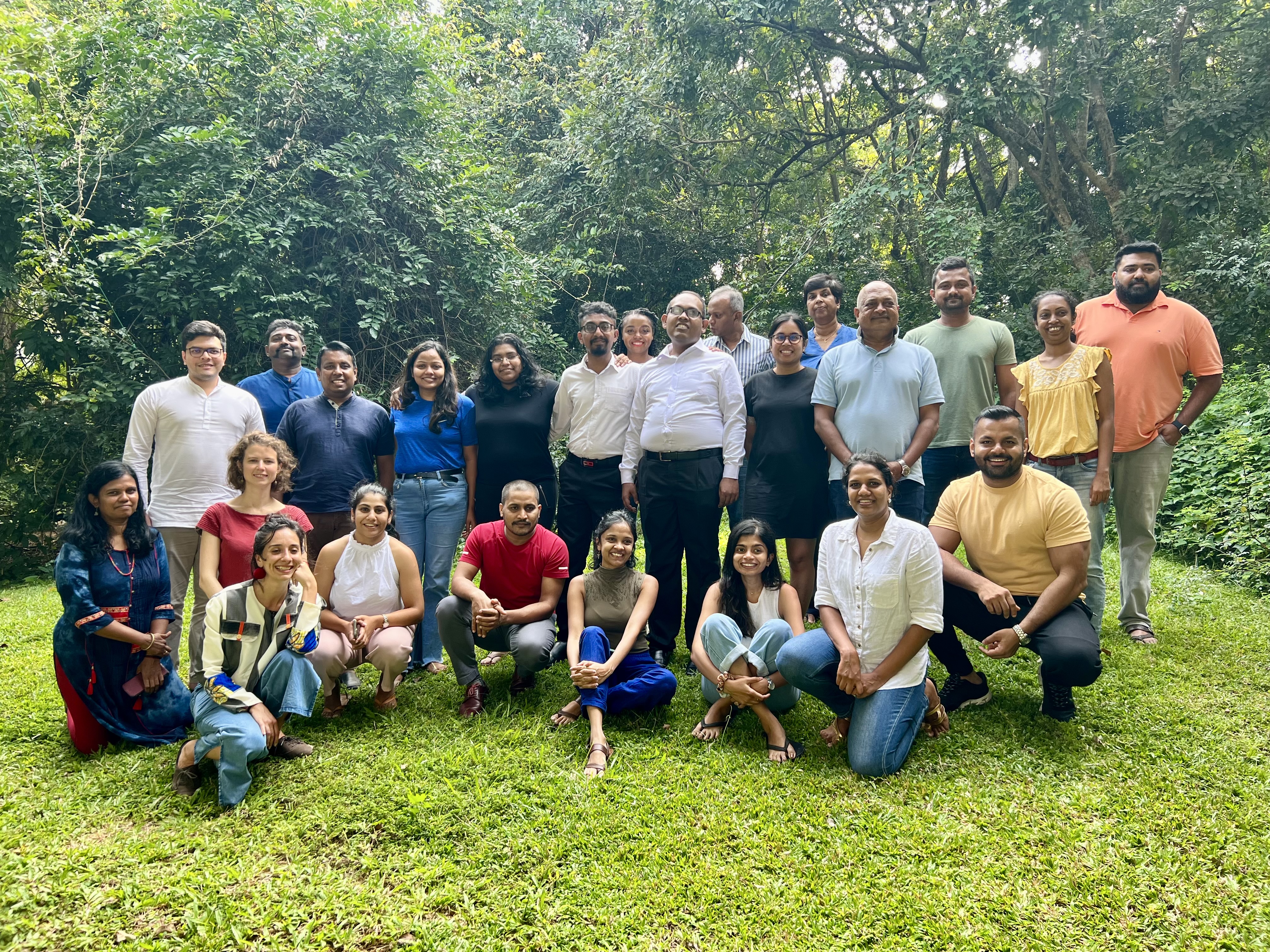 A group of people posing for a group photo. Some are seated on the grass while others are standing. There are trees seen in the background. 
