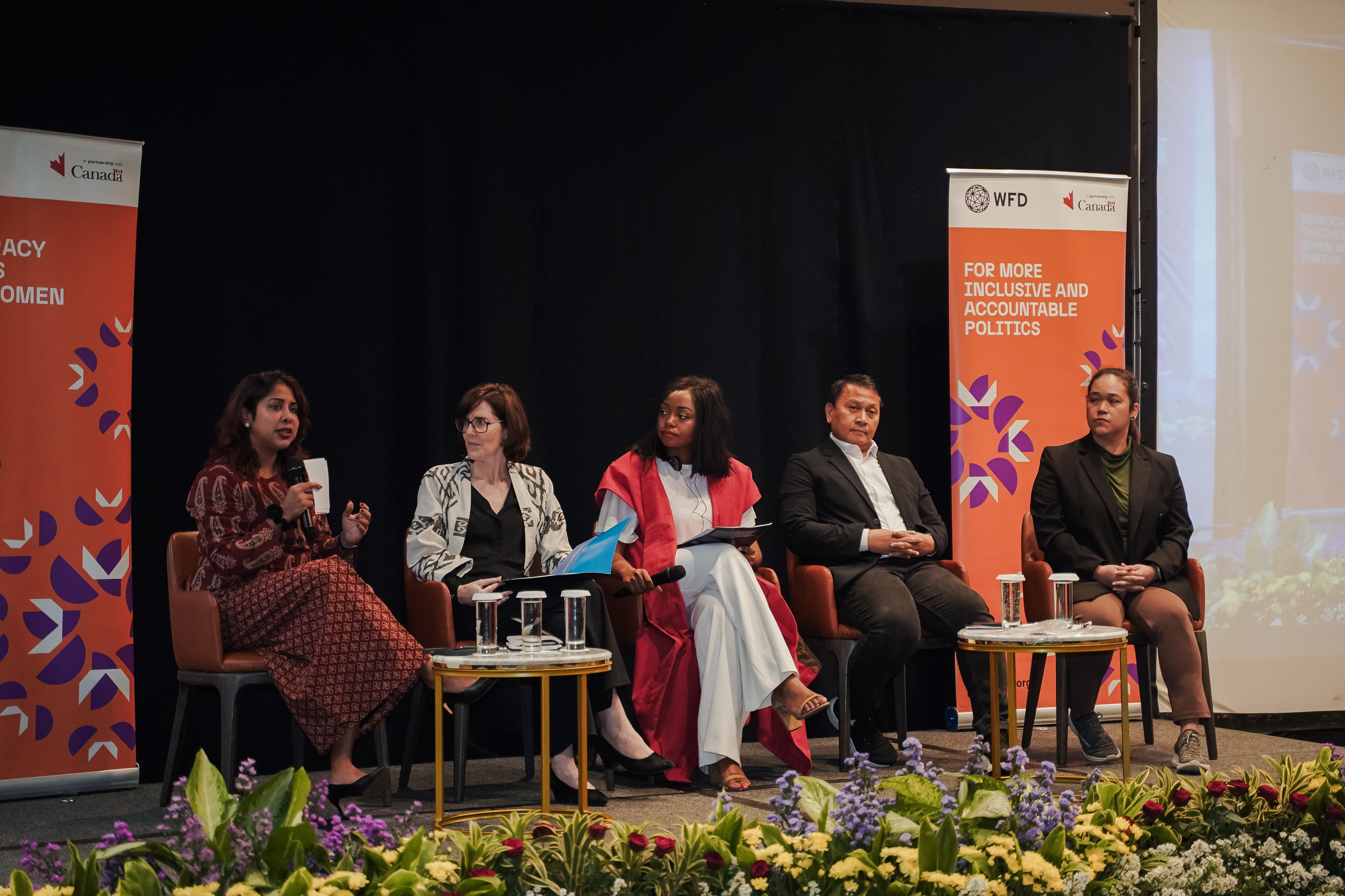 Four women and a man are seated on chairs on a raised platform. A woman in a red dress is speaking into a microphone while others are listening to her. 