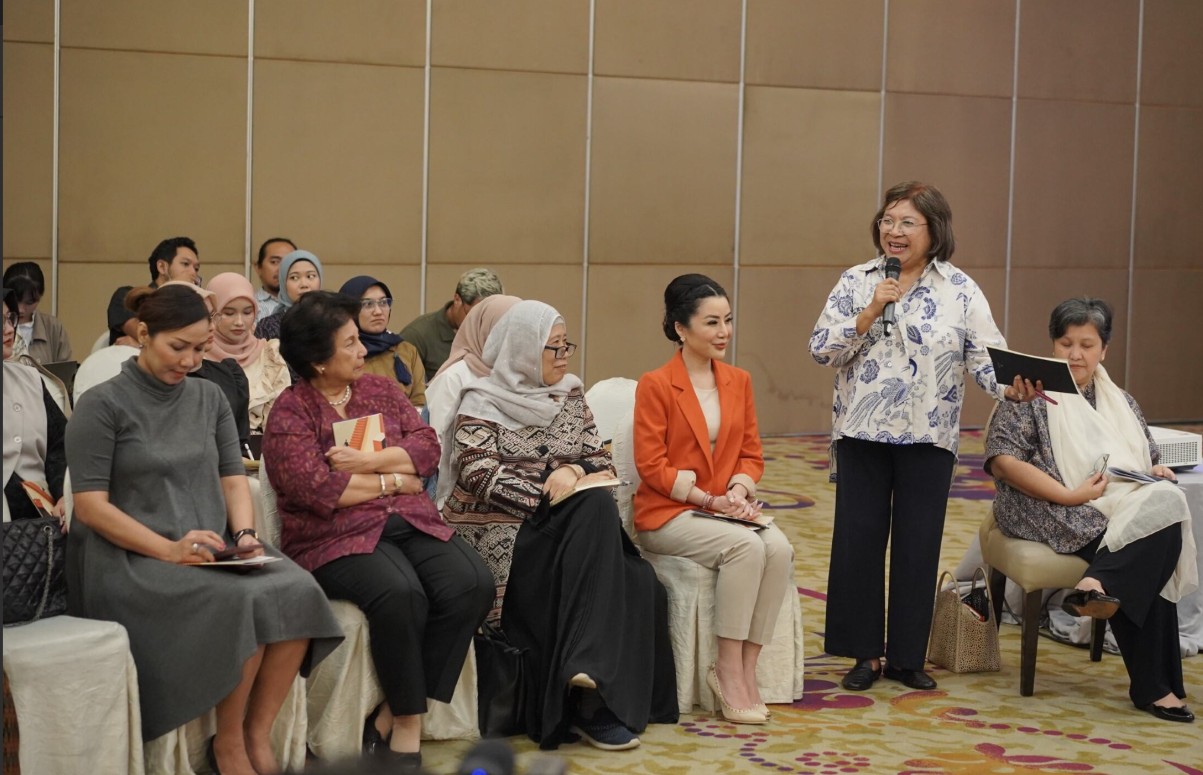 A group of women - each holding a book in their hands - are seated in chairs, while an older woman stands next to them speaking into a microphone. 