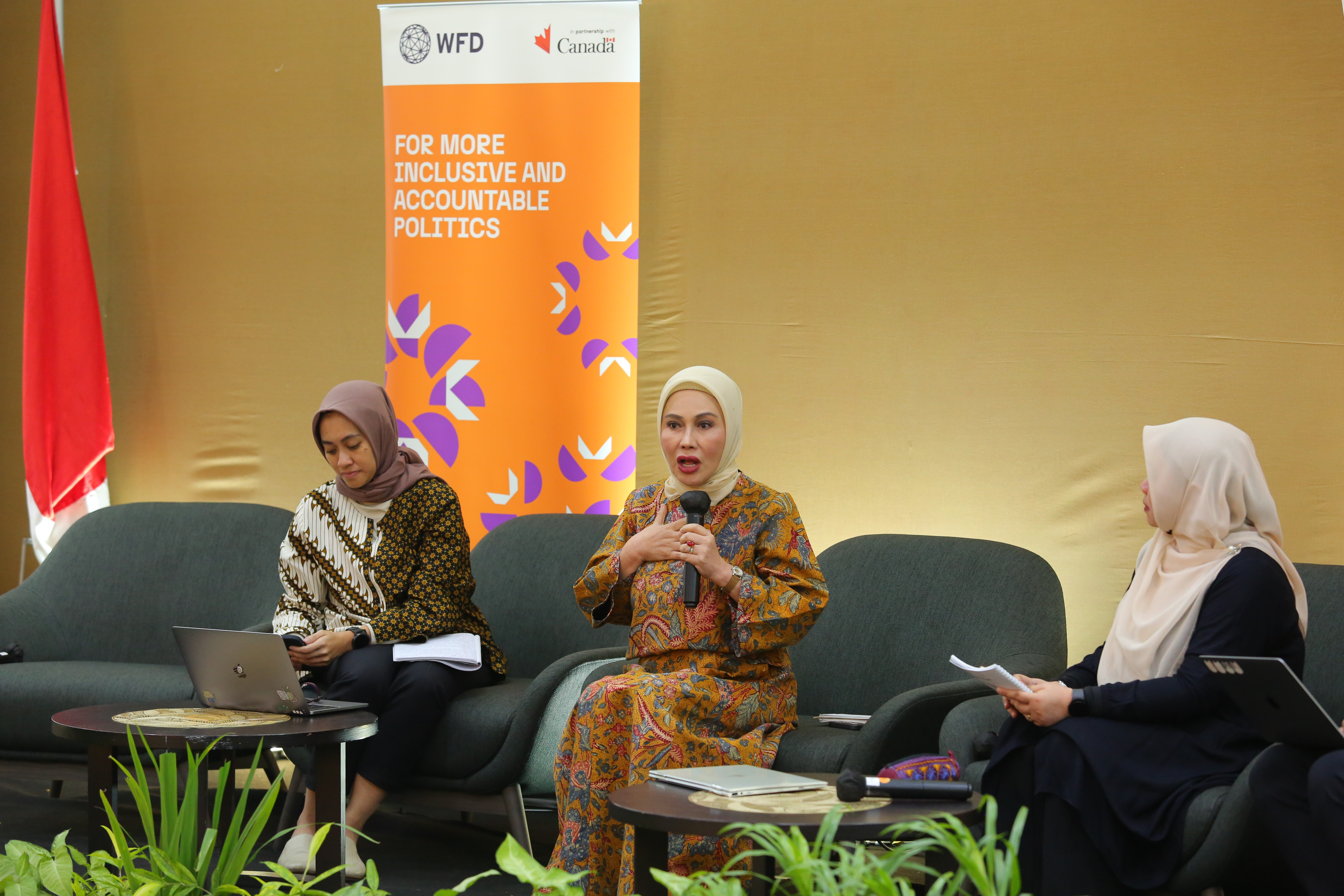 The image shows three women in hijabs sitting on a couch at a seminar. One woman is speaking into a hand-held mic. 