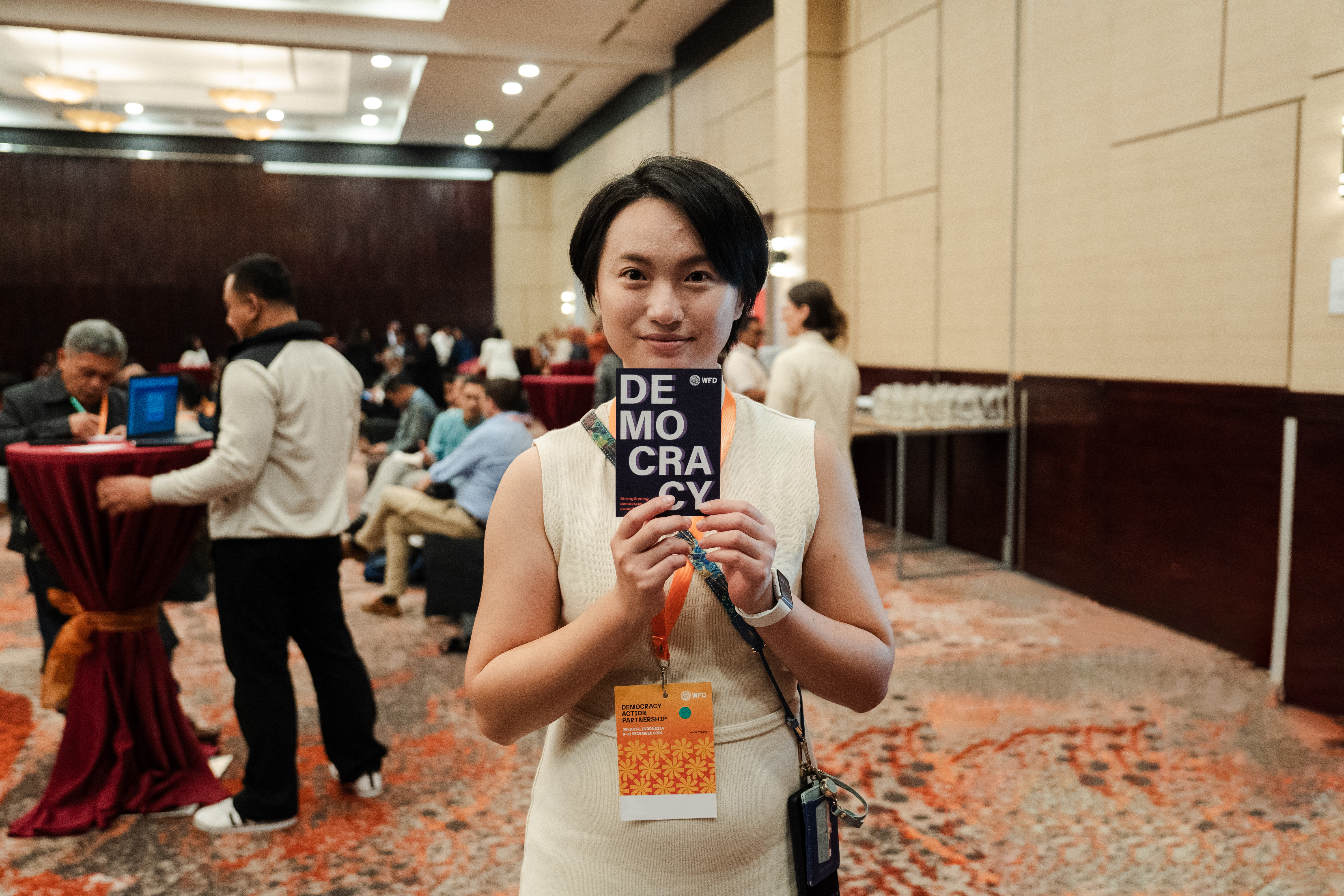 A woman holds up a postcard saying democracy, people at an event can be seen in the background.