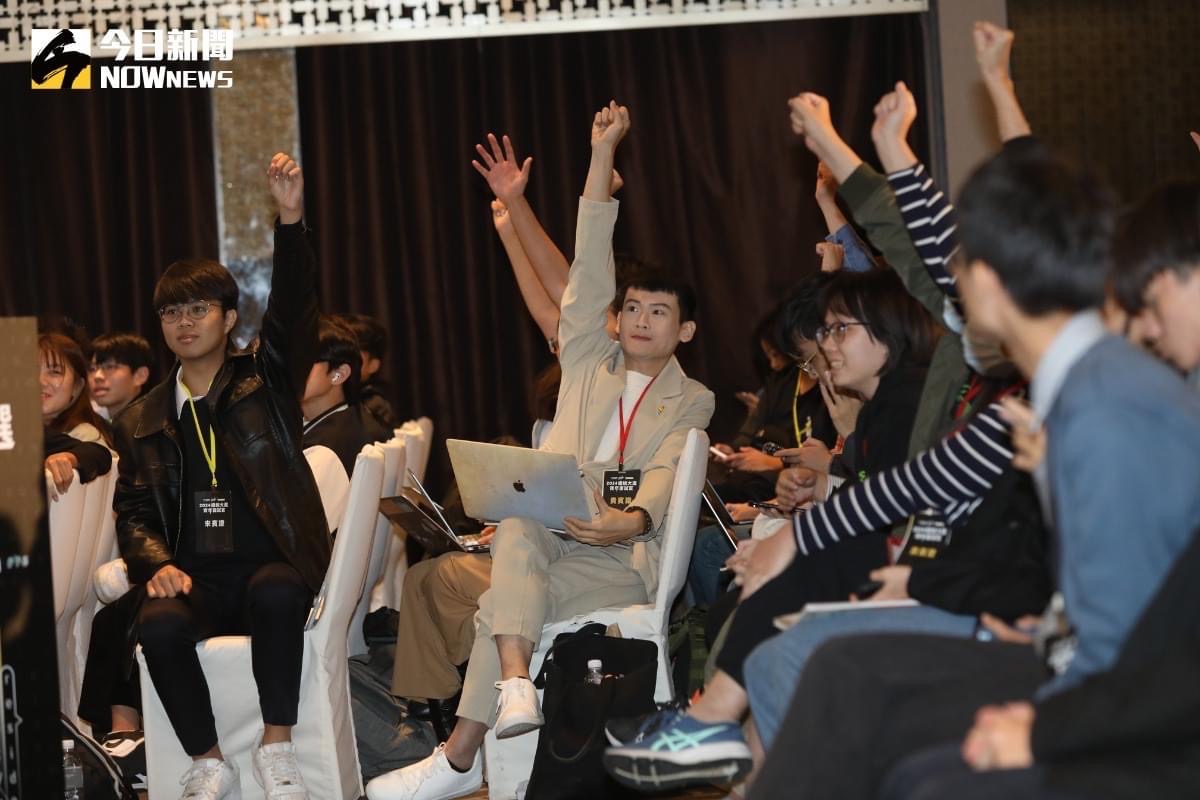 A group of young people are seated in a hall. Some of them are raising their hands and making fists.