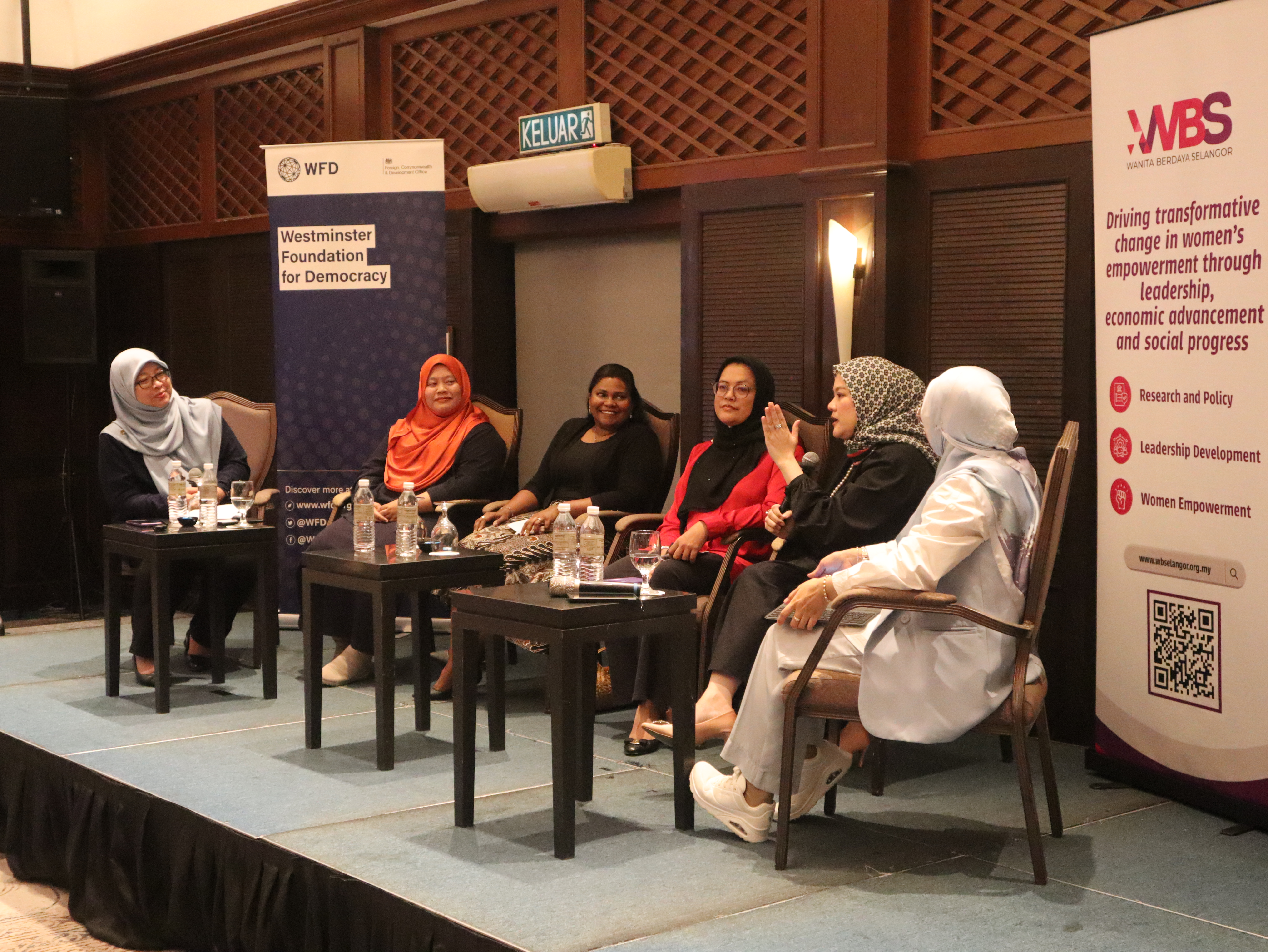 Six women are seated on a stage. One of them is speaking while others seem to be listening to her. There are side tables with water bottles in front of them. 