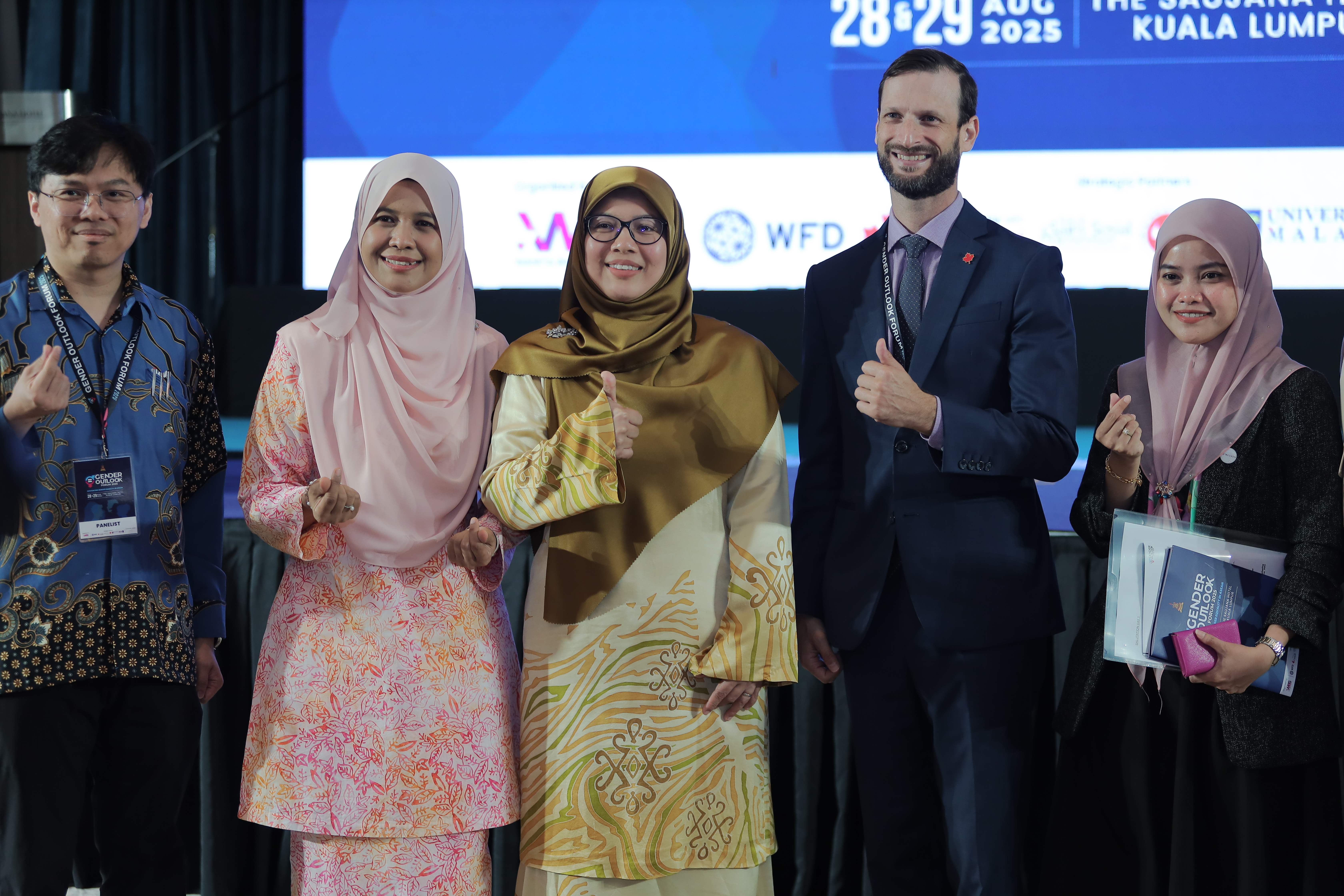 Two men and three women stand next to each other, posing for a photo. They're giving thumbs-up and heart signs with their hands. 
