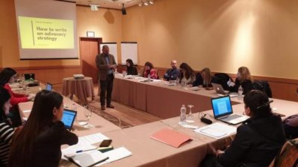 A person speaking at a conference, people sitting at a table around them