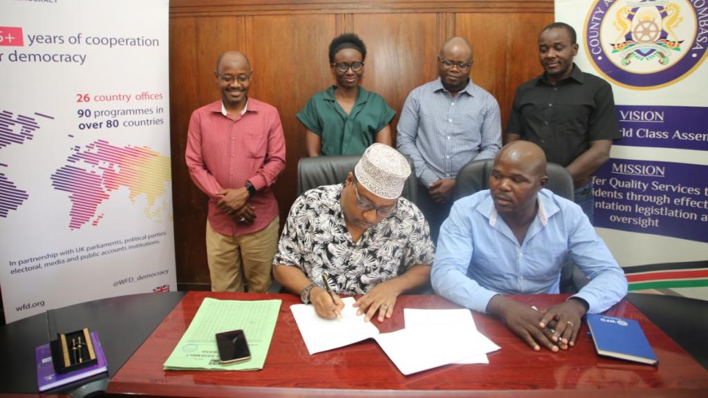 Two people sitting signing an MoU while three are standing behind them