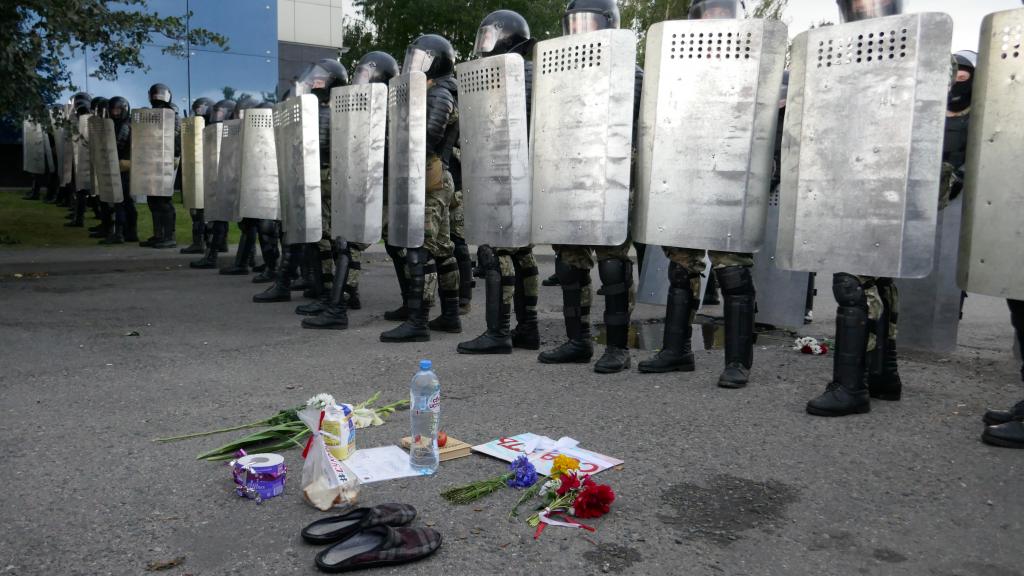 A line of people in helmets holding grey shields, with flowers, shoes and notes at their feet