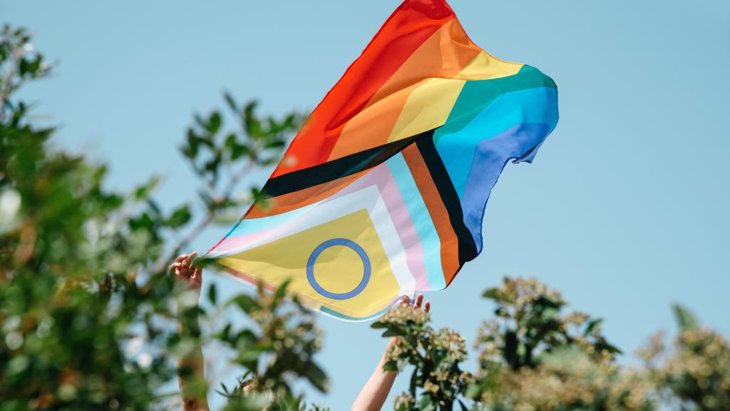 A progressive pride flag being held to fly in the air against a blue sky