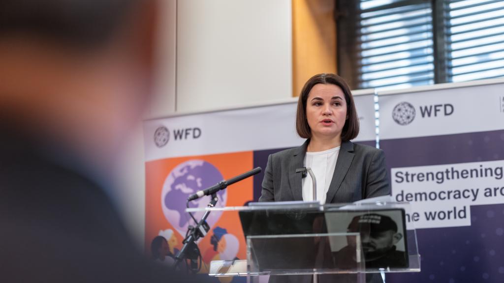 A woman stands at a lectern in front of a microphone, the backs of the audience's heads are in the foreground. She stands next to a sign that says strengthening democracy around the world. The words Inaugural Lecture on the State of Democracy are on a screen next to her.