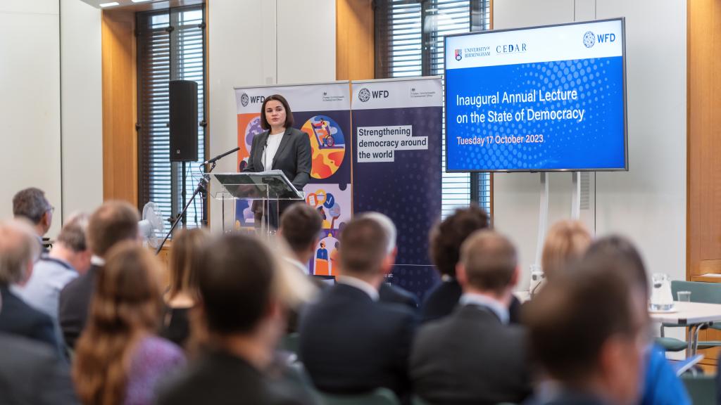 A woman stands at a lectern in front of a microphone, the backs of the audience's heads are in the foreground. She stands next to a sign that says strengthening democracy around the world. The words Inaugural Lecture on the State of Democracy are on a screen next to her.