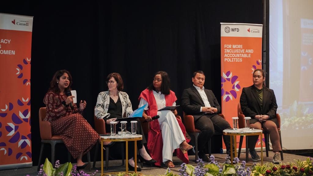 Four women and a man are seated on chairs on a raised platform. A woman in a red dress is speaking into a microphone while others are listening to her. 