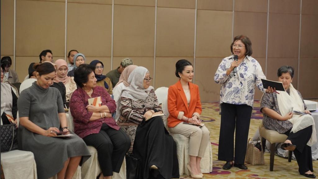 A group of women - each holding a book in their hands - are seated in chairs, while an older woman stands next to them speaking into a microphone. 