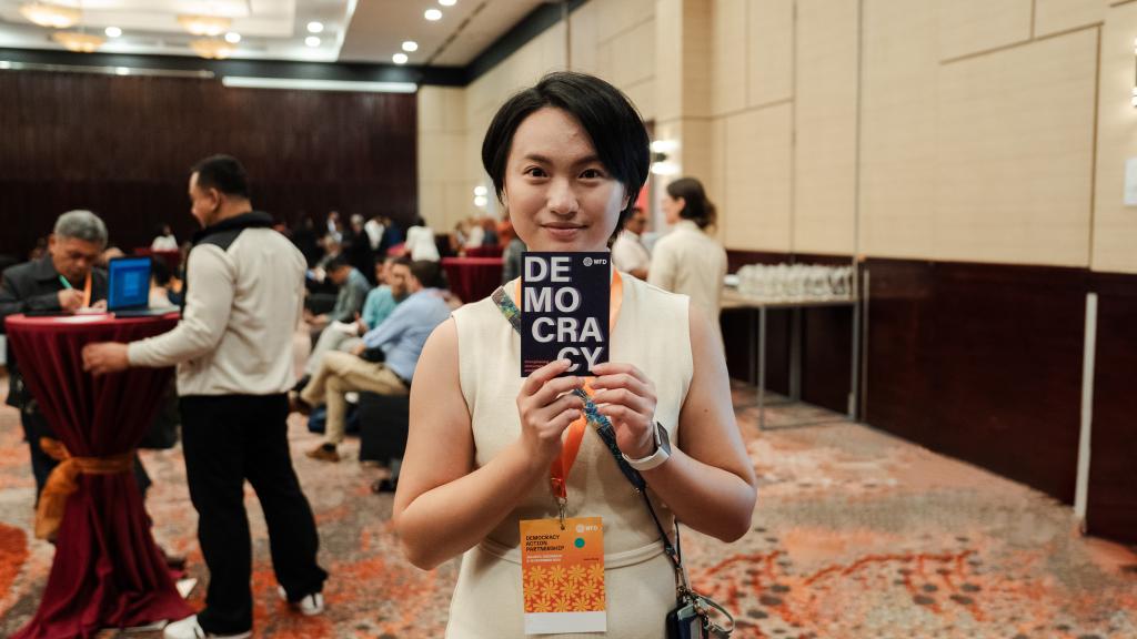 A woman holds up a postcard saying democracy, people at an event can be seen in the background.