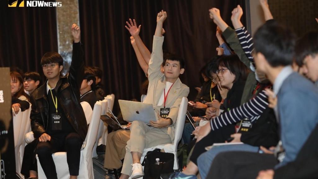A group of young people are seated in a hall. Some of them are raising their hands and making fists.
