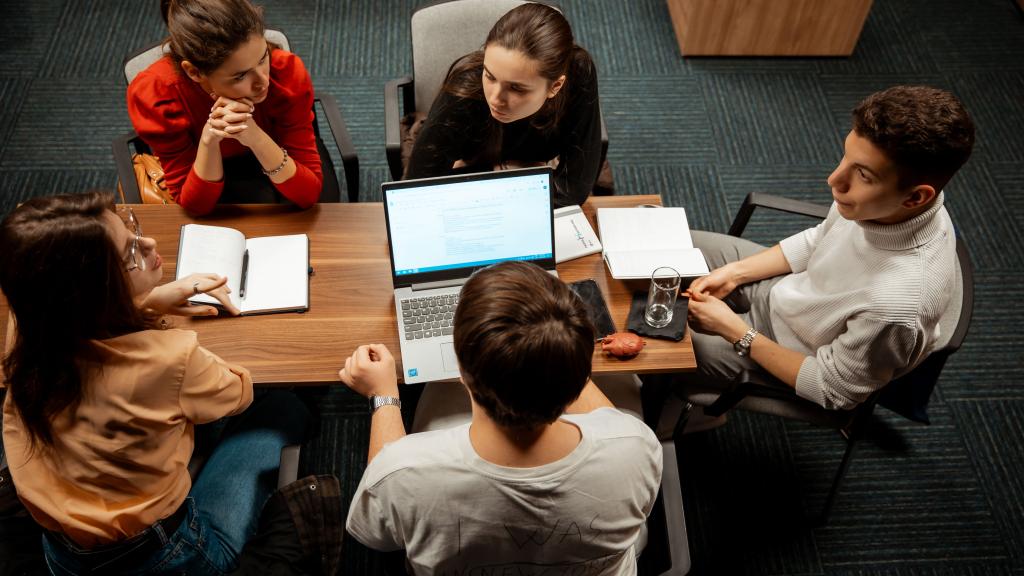 Young participants engaged in a discussion around a desk, with notebooks and a computer on the table.