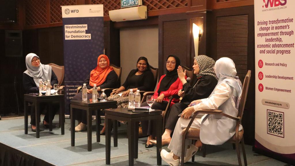 Six women are seated on a stage. One of them is speaking while others seem to be listening to her. There are side tables with water bottles in front of them. 