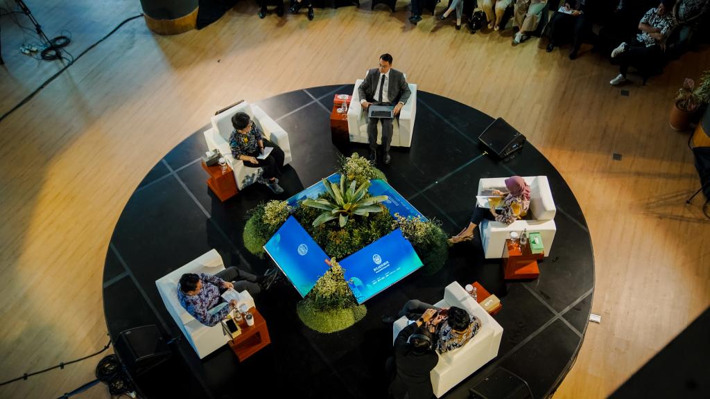 An aerial shot shows five chairs arranged around screens with men and women seated. An audience can be seen in the background.