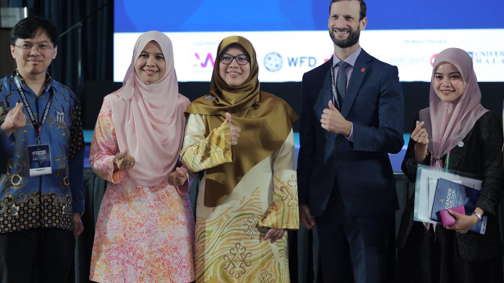 Two men and three women stand next to each other, posing for a photo. They're giving thumbs-up and heart signs with their hands. 