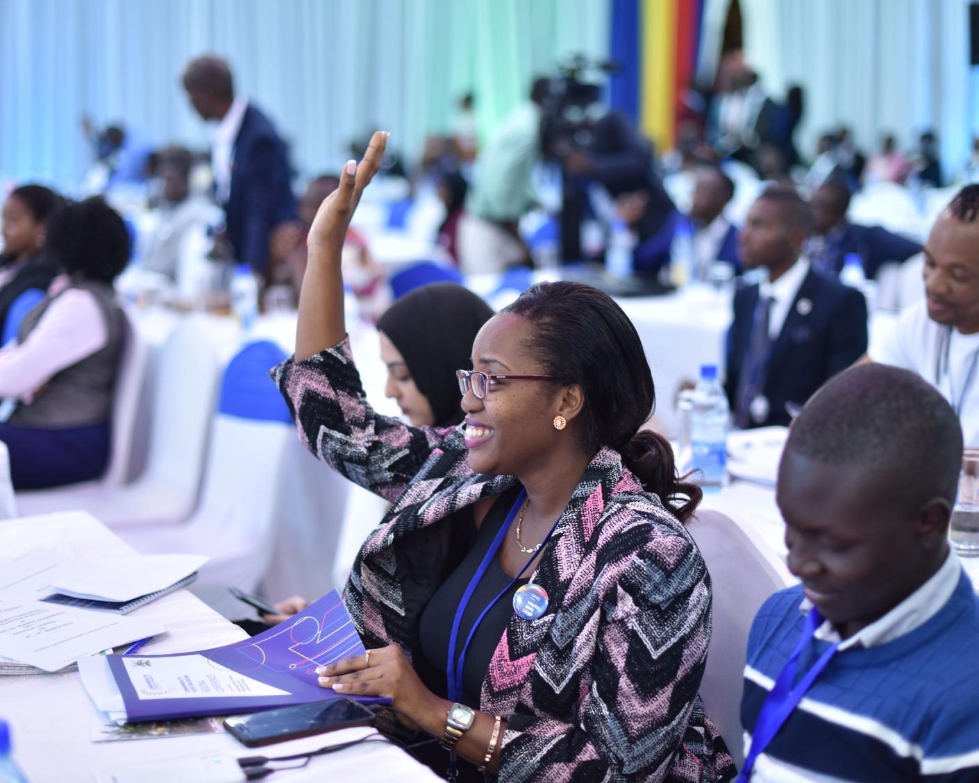 A young woman sitting at a table among other young people from Africa raises her hand