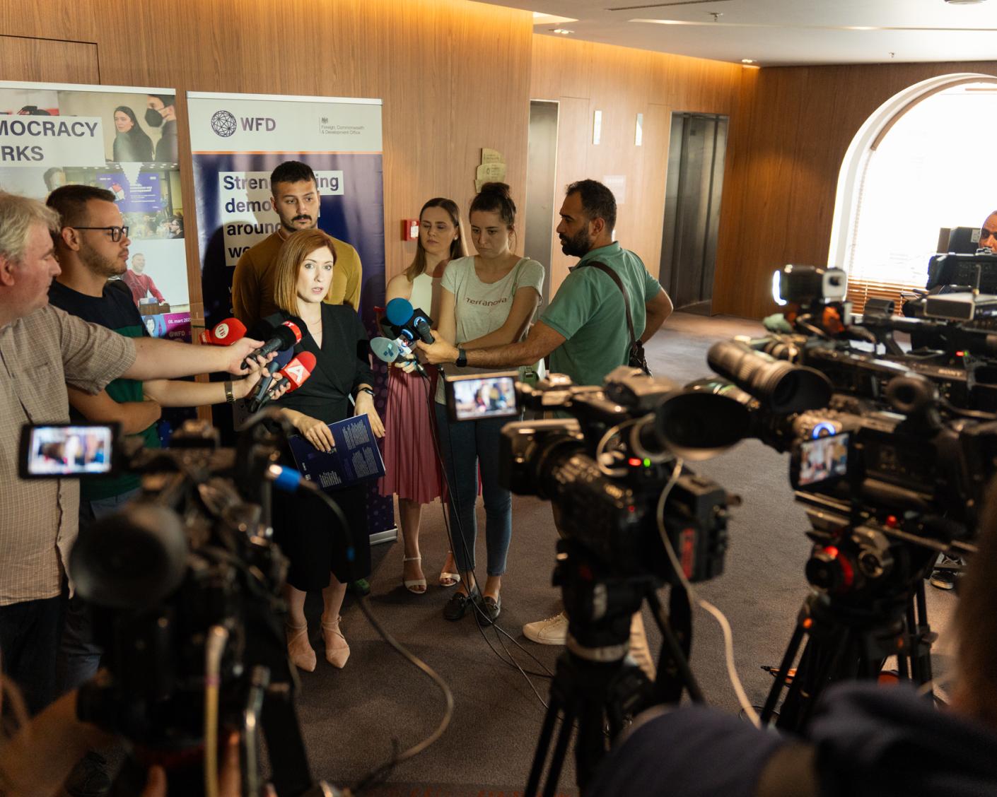 A woman stands in front of WFD banners surrounded by cameras and microphones