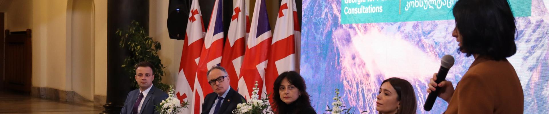 A woman speaks into a microphone at a panel event. In the background are an image of mountains and Georgian flags