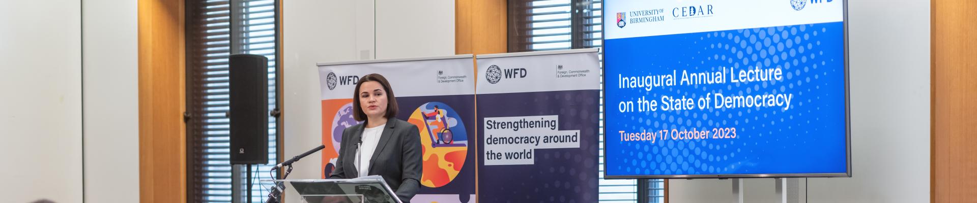 A woman stands at a lectern in front of a microphone, the backs of the audience's heads are in the foreground. She stands next to a sign that says strengthening democracy around the world. The words Inaugural Lecture on the State of Democracy are on a screen next to her.