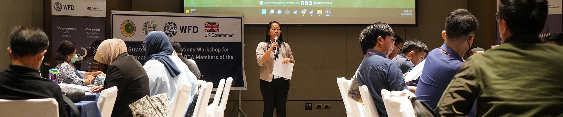 A woman with a microphone stands in front of an audience. Behind her a screen says Bangsamoro Parliament