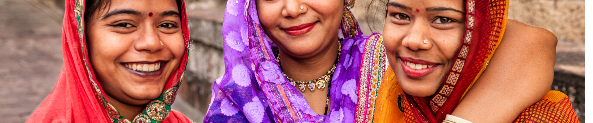 Three women wearing a traditional dress in red, purple and dark orange standing and smiling at the camera  