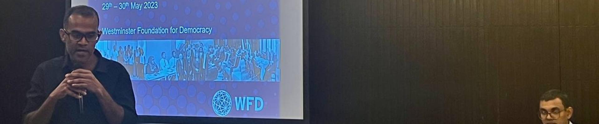 A speaker addressing the audience in a seminar, with a background of WFD slide being projected on the screen 