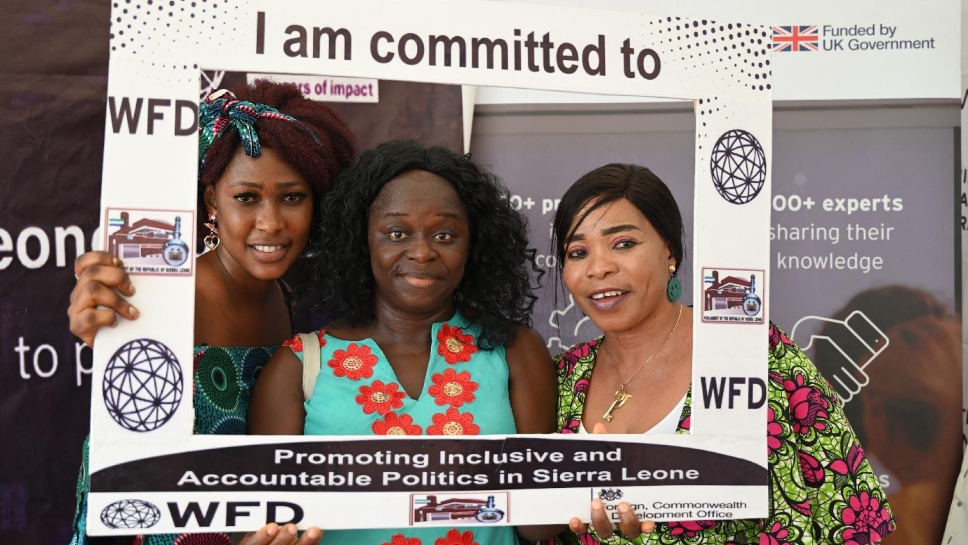 Three women holding up a sign that says I am committed to promoting inclusive and accountable politics in Sierra Leone