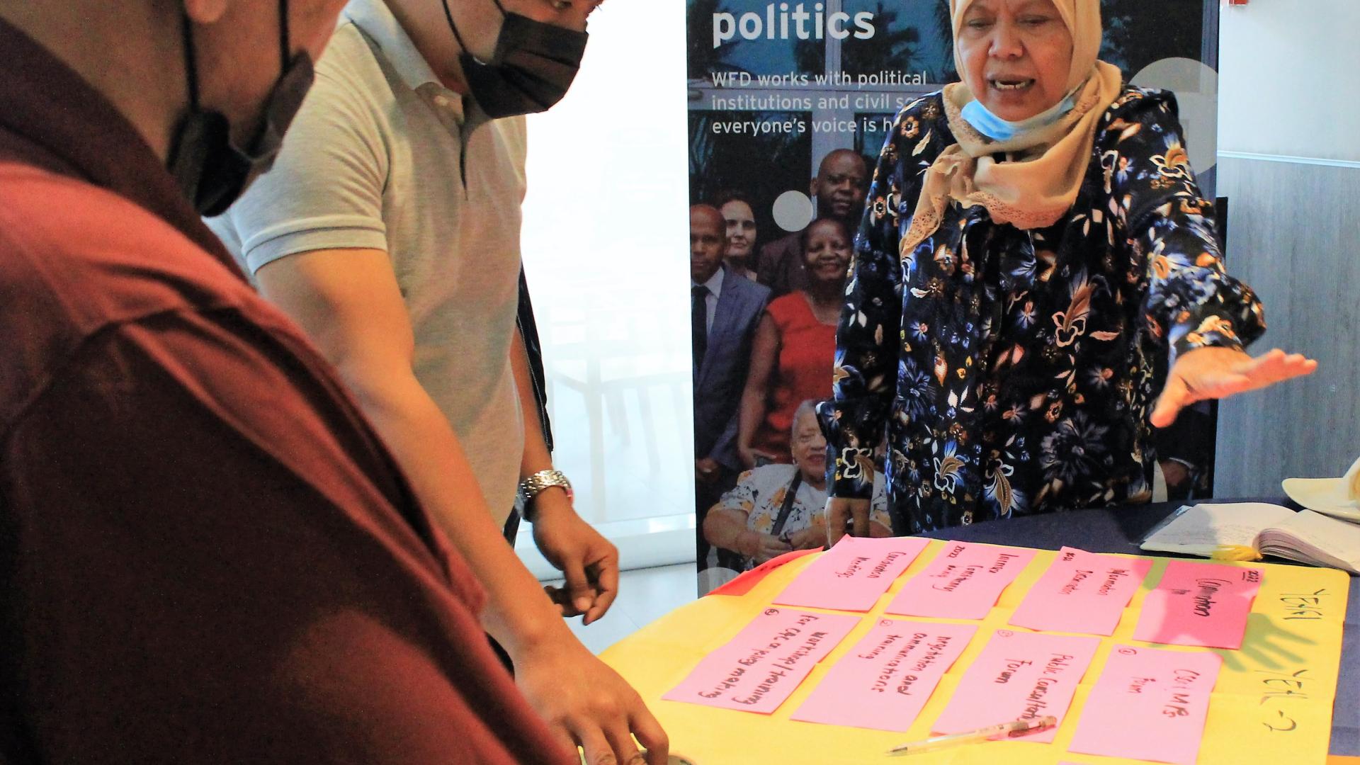 A woman wearing a headscarf stands speaking in front of a table of post-it-notes. Two men in face masks listen with concentrated expressions