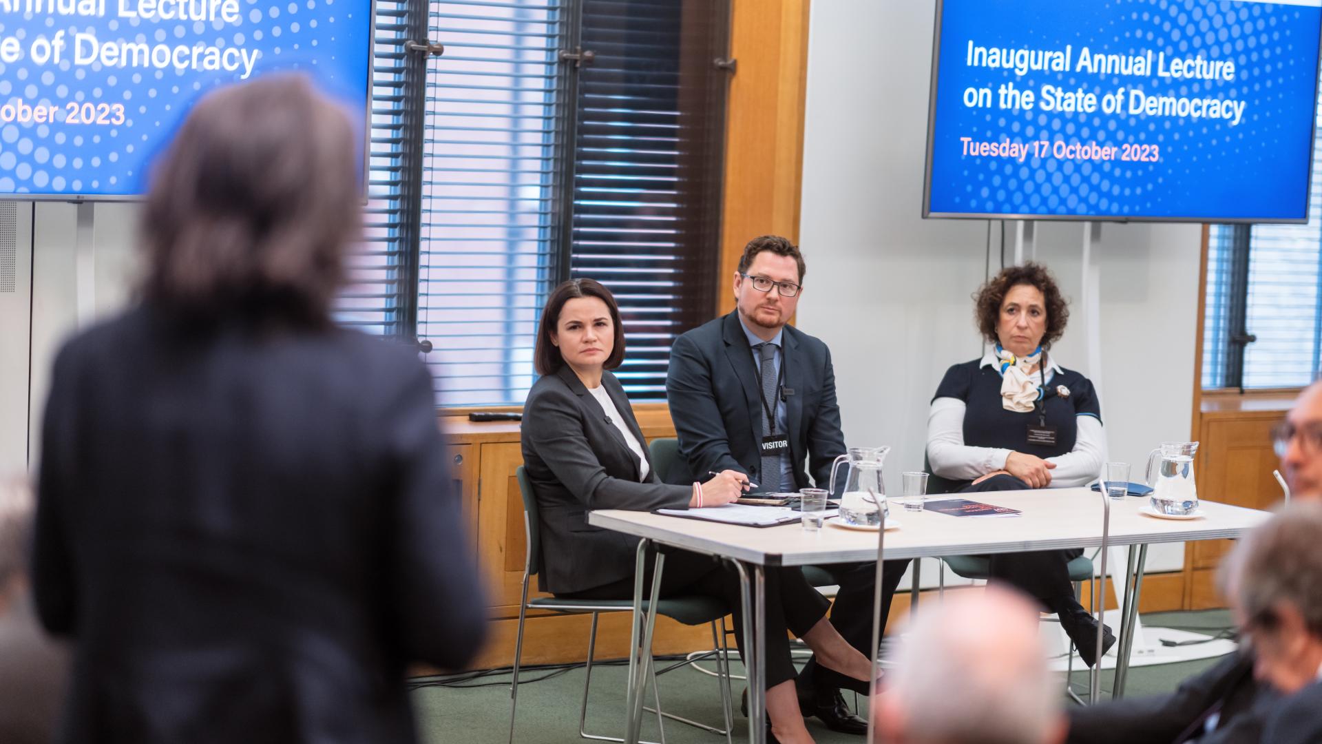 A person in the foreground stands and asks a question to a panel of three seated people