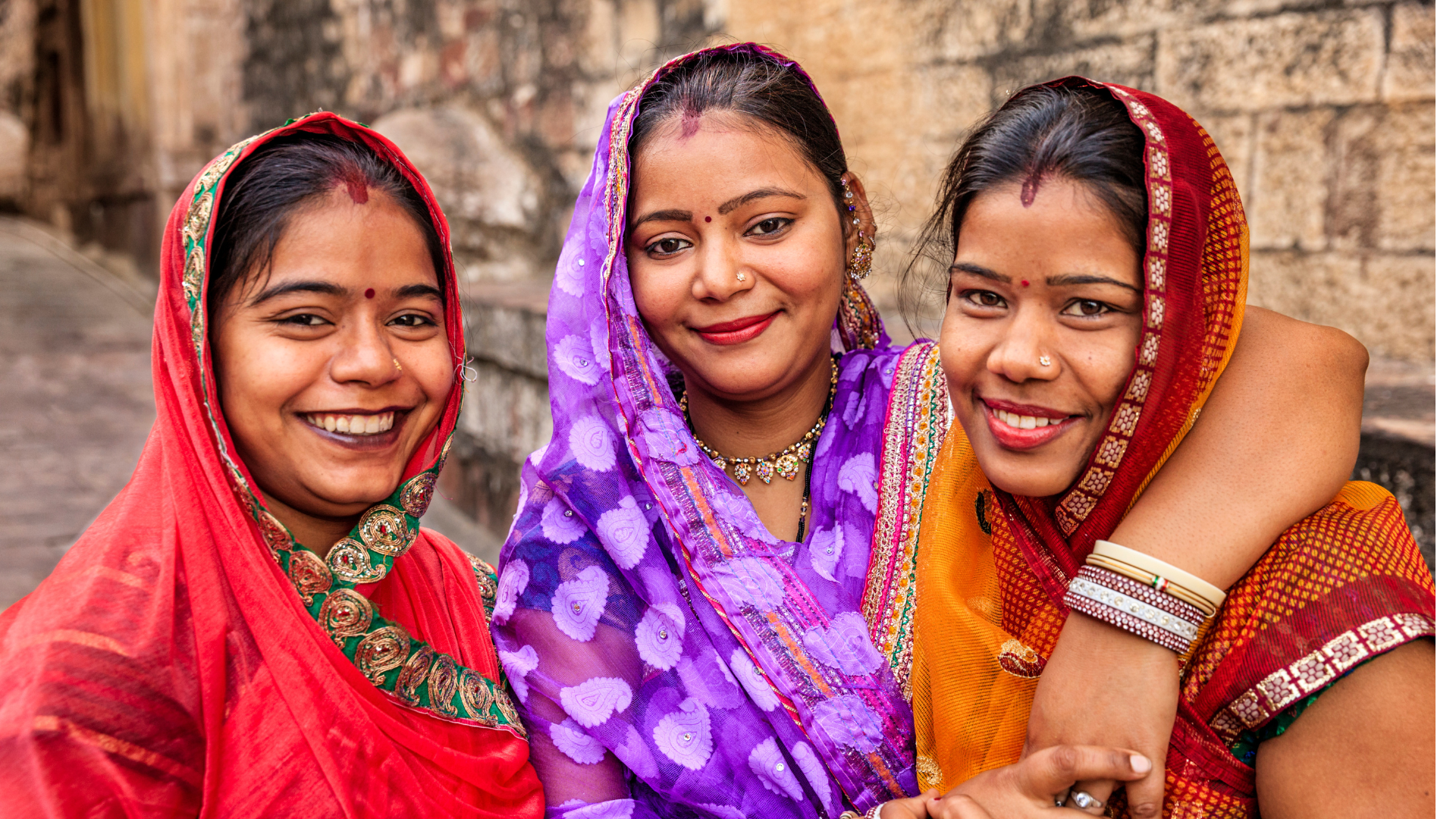 Three women wearing a traditional dress in red, purple and dark orange standing and smiling at the camera  