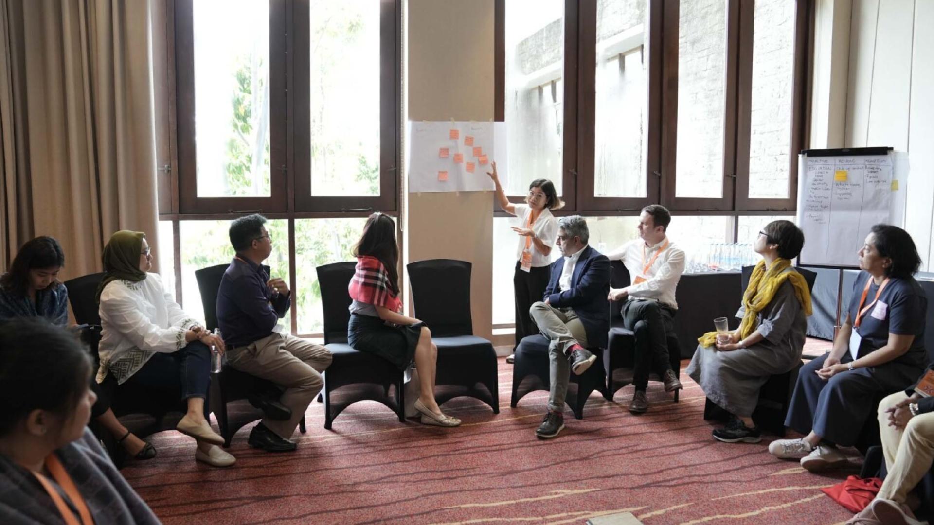 A group of people look to a woman who is presenting a sheet of paper with post-it notes on