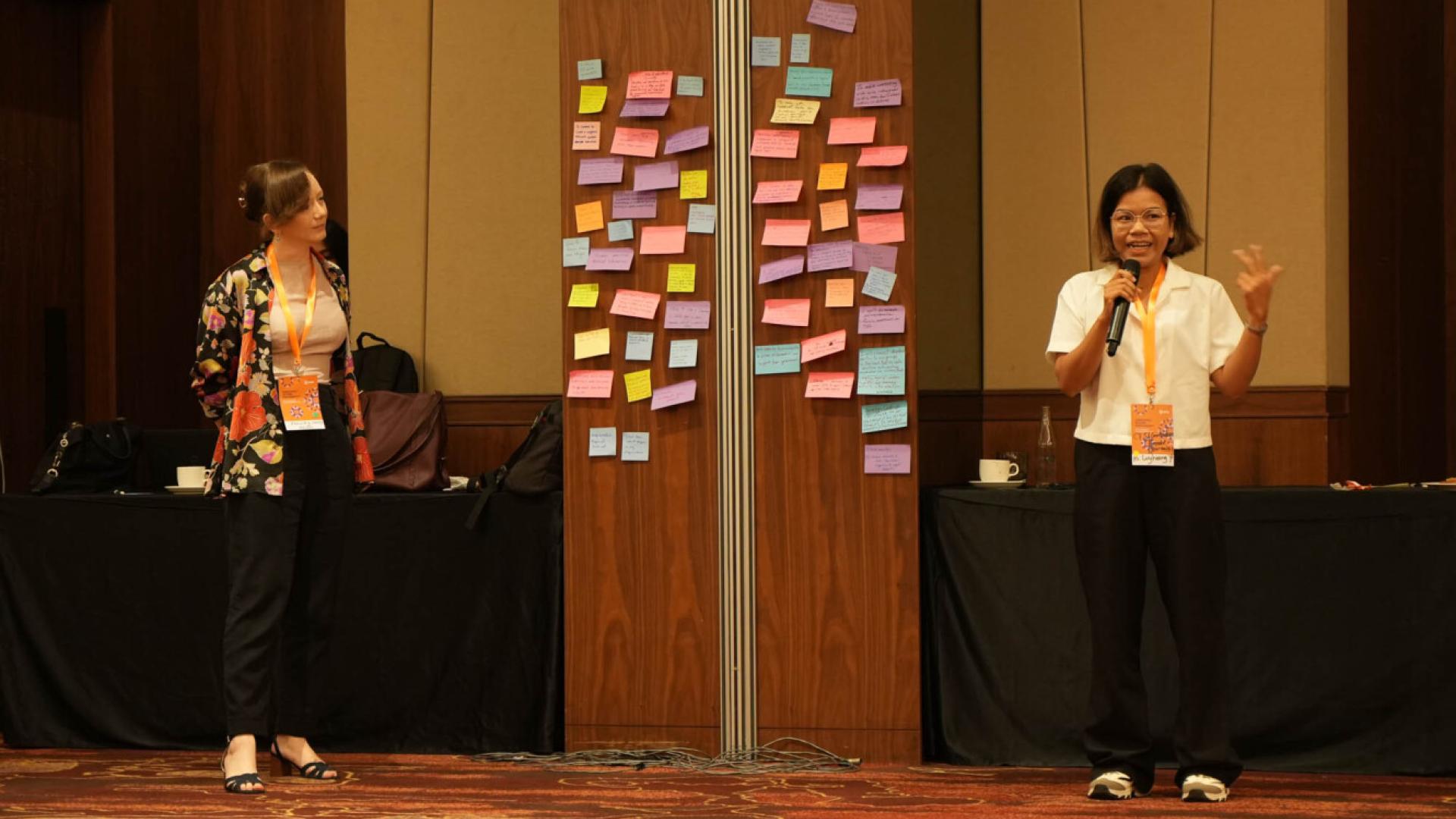 Two women stand either side of a wall of post-it notes, one is speaking into a microphone
