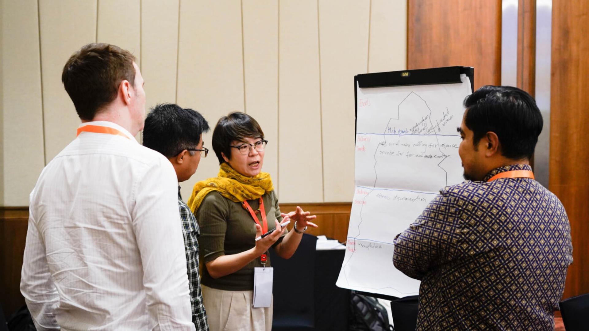 A group of people stand around a flipchat, they are listening to a woman who gesticulates