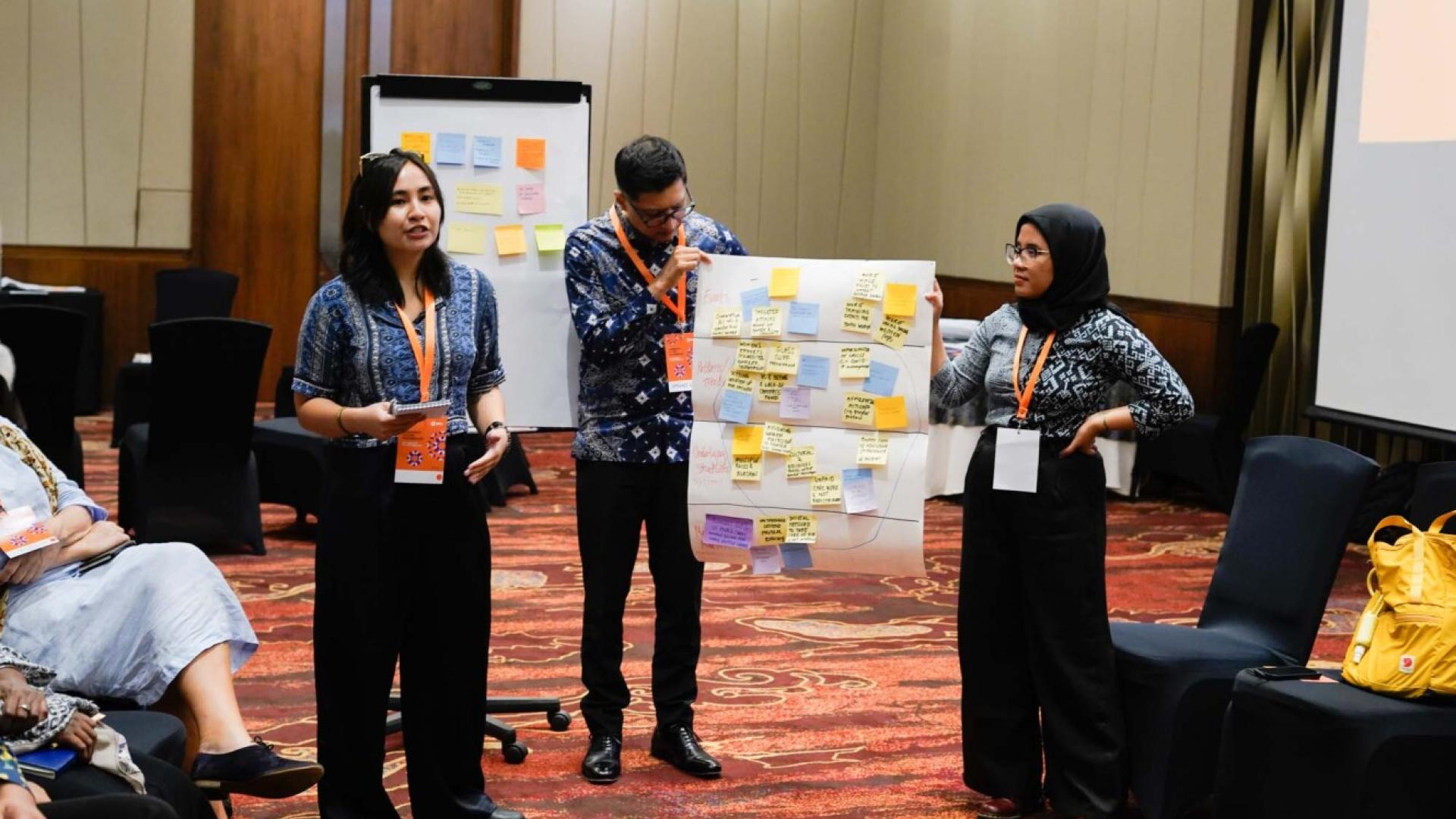 Two people hold up a sheet of paper covered in post-it notes whilst another talks