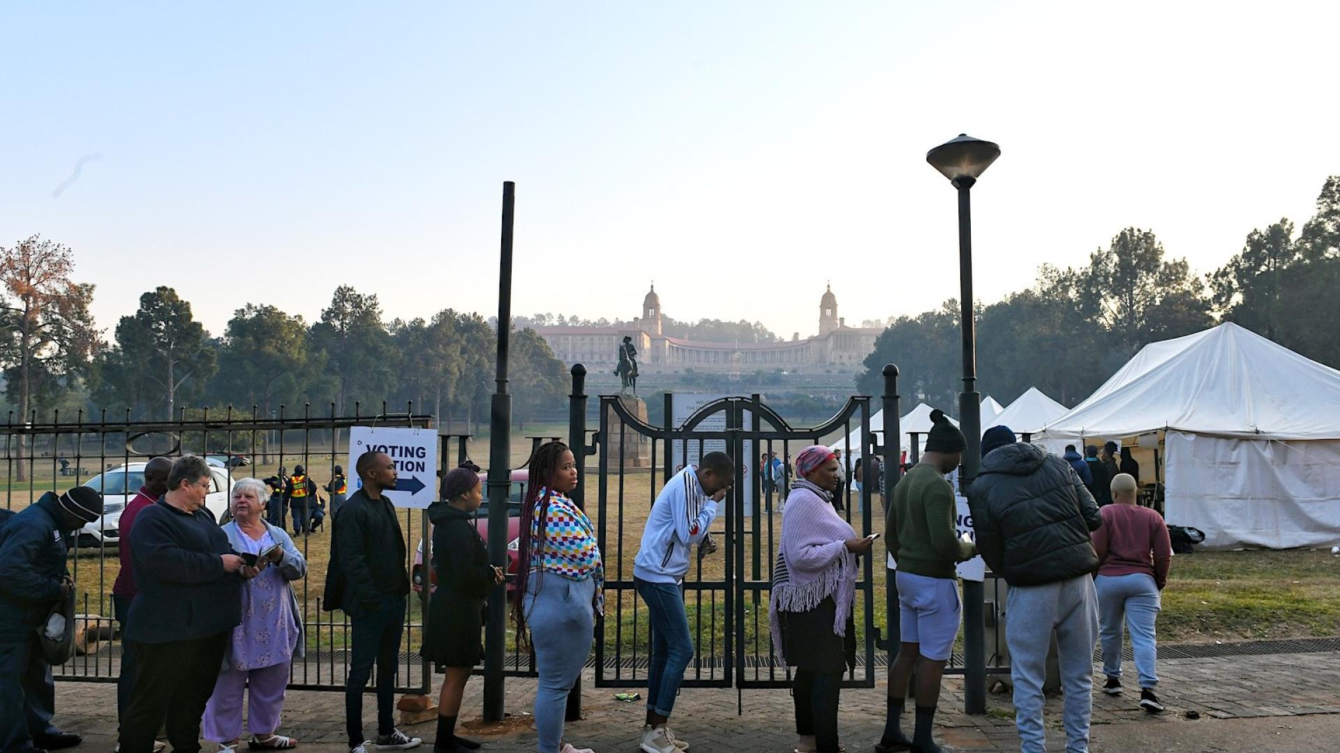 A queue of people standing in line in front of a fence with a polling station sign attached to it.